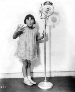 A young girl in a frilly dress stands in front of an old-fashioned NBC microphone, holding up one hand and making a surprised or playful expression.