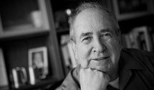 An older man with thinning hair smiles gently, chin resting on his hand, against blurred bookshelves and framed photos in black and white.