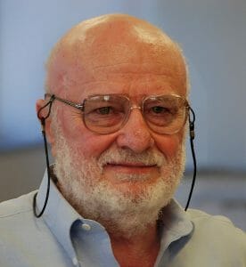 An older man with a bald head, white beard, and glasses with a strap in a light blue shirt looks to the side in soft indoor light.