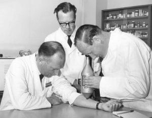 Three men in lab coats, one holding a device against another’s forearm while the third observes. Shelves with bottles and laboratory equipment are visible in the background.