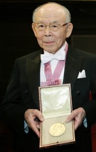 An elderly man in a tuxedo with a pink ribbon around his neck holds an open case with a gold medal, standing indoors facing the camera.