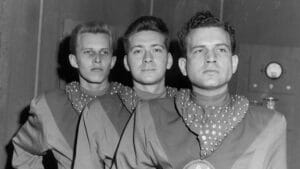 Three men stand side by side in futuristic uniforms with polka-dot scarves, looking serious. The image is black and white, and the background appears to be a sci-fi or retro setting with control panels.