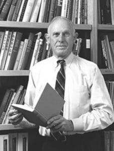 A man in a dress shirt and tie stands by bookshelves, holding an open book and facing the camera.