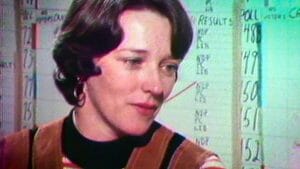 A woman with short brown hair and hoop earrings looks down thoughtfully in front of a whiteboard covered in handwritten election stats.