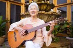 An elderly woman with white hair sits by a fountain, smiling and playing guitar, in light clothes near plants and wood-framed windows.
