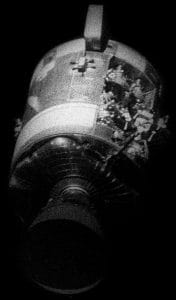 A damaged spacecraft drifts in space against a black backdrop, with exposed wreckage and torn panels revealing inner components.