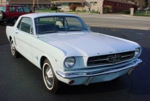 A light blue 1960s Ford Mustang coupe with vintage wheels and chrome accents is parked along a street.