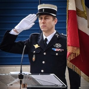 A uniformed officer wearing a hat and white gloves salutes beside a French flag during a formal event, standing behind a clear lectern.