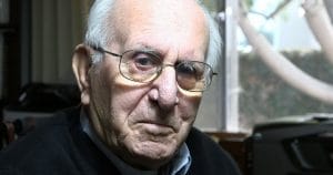 An elderly man with glasses and a serious look sits by a window indoors, wearing a dark sweater. Trees blur in the light outside.