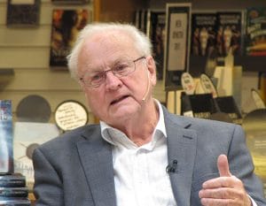 An elderly man with white hair and glasses, wearing a gray suit jacket and white shirt, speaks into a microphone headset while sitting in front of a bookshelf filled with books.