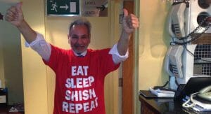 A man smiling and giving two thumbs up in a red "EAT SLEEP SHISH REPEAT" shirt, with telephones and restroom sign behind him.