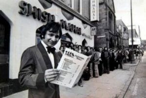 A man in a suit and bow tie stands outside a restaurant, holding up a menu and smiling; people line up along the sidewalk.