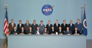 Nineteen men in suits pose formally by a NASA logo, seated and standing at a long table with US and NASA flags on each side.