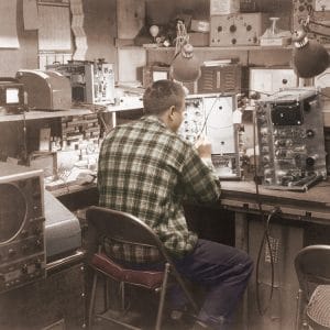 A man in a plaid shirt sits at a cluttered workbench filled with electronic equipment and tools, appearing to repair or assemble a device in a vintage-looking workshop.