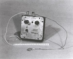 Black and white photo of an early transistor pacemaker device with dials labeled Pace Rate and Output, wires extending from it, and a ruler showing scale in front.