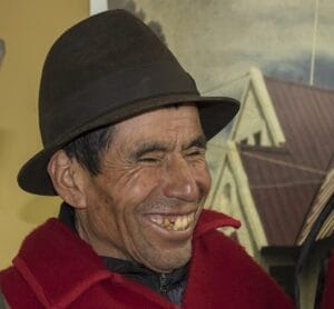 A man in a brown hat and red poncho smiles, showing his teeth. Behind him is a sloped-roof building with muted colors.