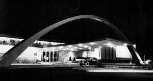 A black and white photo shows a modernist building at night with a big arch, glass entry, vintage car, and soft lighting outside.