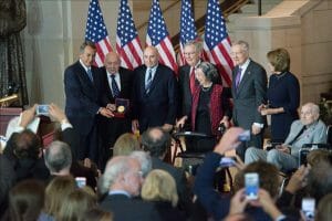 A group, some elderly and in wheelchairs, poses by American flags as a man holds a medal. People take photos with their phones.