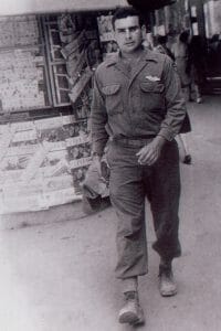 A young man in military uniform walks confidently toward the camera on a city street with magazine stands and people behind him, World War II vintage.
