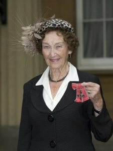 An elderly woman in a black suit and patterned hat smiles, holding an award medal on a red ribbon outside by a building window.