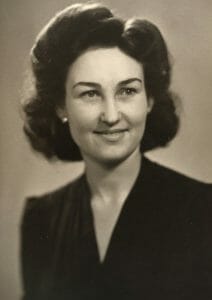 Black and white portrait of a woman with wavy dark hair above her shoulders, in a dark dress, smiling gently and looking left. It appears to be from the 1940s.
