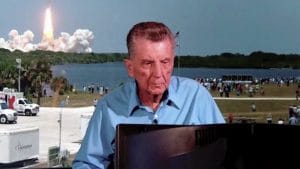 An older man sits at a desk outdoors as a rocket launches across water in the background, with a crowd watching the event from the midground.