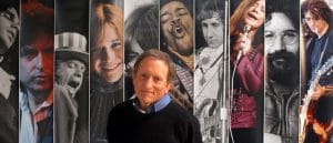 A man stands smiling in front of a mural with portraits of 1960s rock musicians singing and playing instruments.