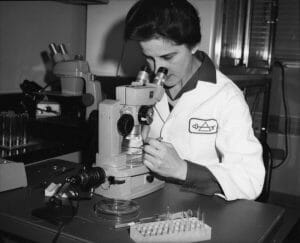 A woman in a lab coat uses a microscope at a lab desk with test tubes, petri dishes, and equipment, focused on examining a sample.