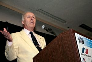 An older man in a yellow suit speaks at a lectern with a microphone during a conference. Logos for ISDC 2001 and National Space Society are visible.