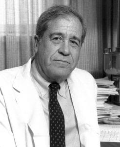 A grayscale photo of an older man in a white coat and patterned tie, sitting in an office with stacks of papers and files visible behind him. The man has short gray hair and a serious expression.
