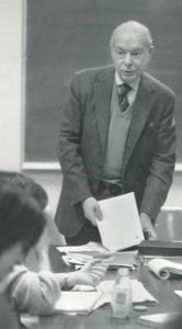 An older man in a suit stands at the front of a college classroom, holding papers and speaking to students seated at a table. A chalkboard is visible in the background.
