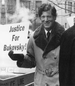 A person in a double-breasted coat and gloves holds a sign reading Justice For Bukovsky! while smiling at a protest, with police tape visible in the foreground.