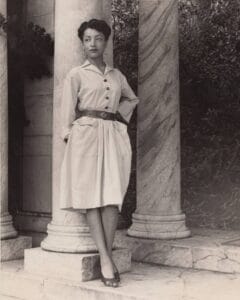 A woman in a belted shirtdress and open-toe shoes stands against a marble column outdoors, with her hands in her pockets and trees in the background. It is a black and white period photo from the 1940s.