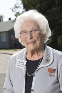 An elderly woman with white hair and glasses smiles outside, wearing a grey blazer with medals and a “Betty Webb” badge, house behind.