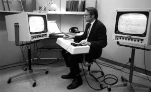 A man in a suit sits at a vintage dual-monitor computer workstation, holding a pointing device amid mid-century office equipment.