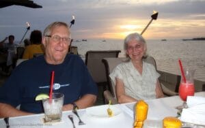 A smiling man and woman sit at a seaside restaurant during sunset, enjoying drinks and food laid out on their table. In the background, torches glow and boats rest on the water.