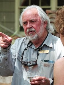 An older man with white hair and a beard, wearing a light gray shirt, name badge, and glasses around his neck, gestures while speaking to another person outdoors.