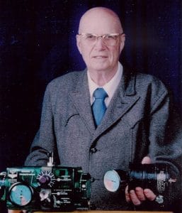 An older man wearing glasses, a grey suit, and a blue tie stands holding respirators with dials and gauges against a dark background.