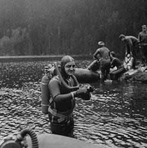 A scuba diver in an old-fashioned wetsuit and gear stands in shallow water, holding equipment, while several other divers and people gather near inflatable rafts on the shore in the background.