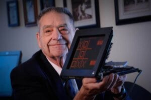 An elderly man in a suit smiles, holding an early computer displaying tic-tac-toe; framed photos are visible on the wall behind him.