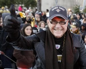 A smiling man in a black leather jacket and WHO DAT Saints cap gives a thumbs-up at an outdoor event with fans in black and gold.