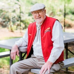 Elderly man with white beard and glasses sits on a picnic bench outdoors, wearing a cap and an embroidered red vest; sunlit green background.