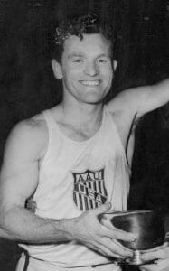 A smiling male athlete in a sleeveless top with an AAU USA emblem holds a trophy bowl in his right hand.
