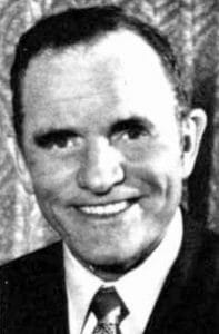 Black and white photo of a man in a suit and tie, smiling at the camera with a patterned background.