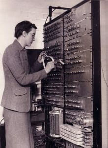 A woman wearing a suit operates an early computer, loading paper tape onto a tall computer panel in a vintage lab setting.