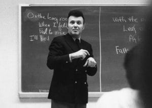 A man in a dark suit stands in front of a chalkboard, gesturing with his hands. The chalkboard behind him has handwritten text.