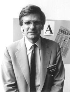Brainerd in a suit and tie, wearing glasses and a name badge, stands in front of a wall with large printed letters and a newsletter page in the background.