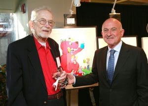 Two men in suits smile in front of colorful Clifford the Big Red Dog artwork; one holds wine at what looks like an art event.