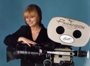 A woman with reddish hair in a black blazer smiles while posing by a large vintage Panavision film camera, blue background.