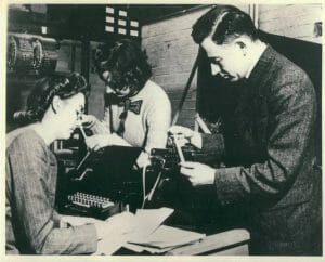 Three people at a desk with an early computer. One woman sits in front of a keyboard computer terminal while another in the background looks at output on a strip of paper. A man stands beside them also looking at an output strip.